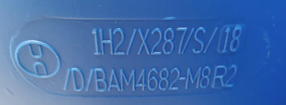 Blue surface with embossed text: "1H2/X287/S/18" and "D/BAM4682-M8R2." Symbol on left. Simple, industrial design.